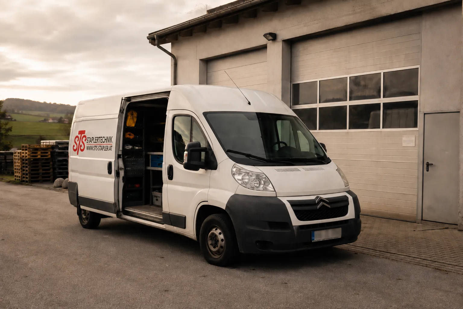 Weißer Servicebus der STS Staplertechnik Salzer mit geöffneter Seitentür vor einer Werkstatthalle in ländlicher Umgebung.